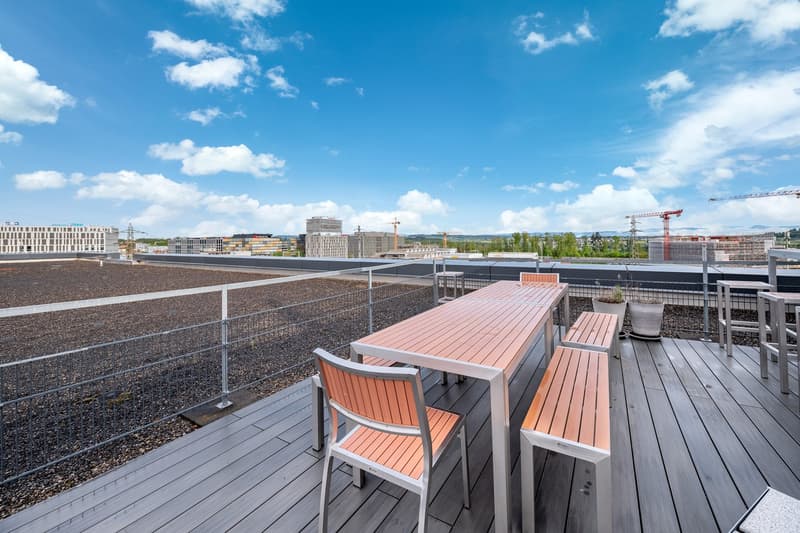 Wooden deck, long outdoor dining table, chairs, benches, potted plants, metal railing, fenced perimeter, panoramic city view