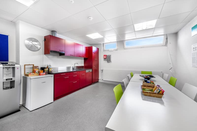 Modern kitchen, red cabinets, white walls, central island with seating, appliances and cooking utensils on countertop, modern dining table and chairs, window providing natural light, industrial aesthetic, and grey flooring