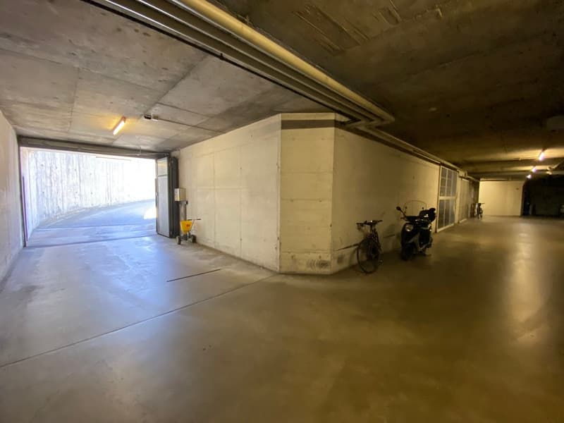 spacious parking garage, with concrete floors, metal pipes, ceiling mounted lights, several parking spots, two bicycles parked, one with a helmet on the seat, a small yellow trolley near the wall, a metal post to the left