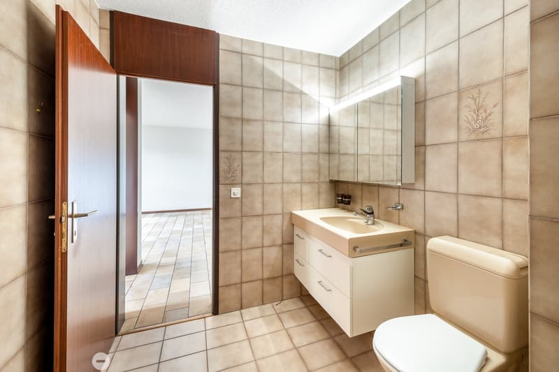 A small bathroom with beige tiles, a sink, a mirror, a toilet, and an open door leading to another room.