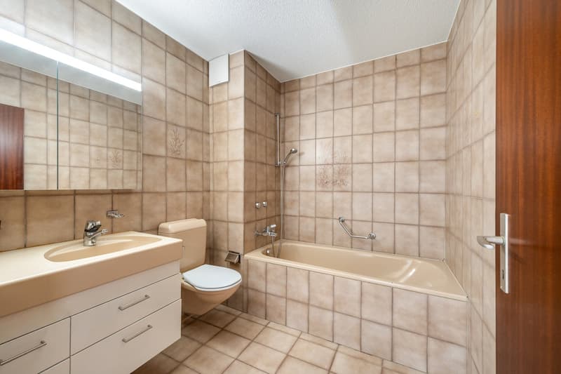A bathroom with bathtub, toilet, and sink. The walls and floor are tiled with beige tiles. There is a mirror on the wall above the sink.