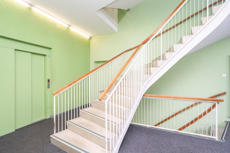 The image shows a staircase with beige steps, white railings, and wooden handrails. An elevator door is on the left side, and a ceiling light is visible on the right. The walls are painted green.