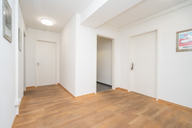 Wide corridor with white walls and wooden flooring. Two closed doors, and one opened leading to a smaller room. A picture and a poster on the wall.