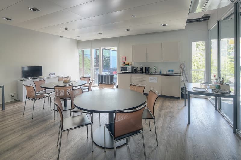 Spacious open-plan office or breakroom with large round dining table, chairs, kitchen area with cabinets and appliances, and large windows providing natural light