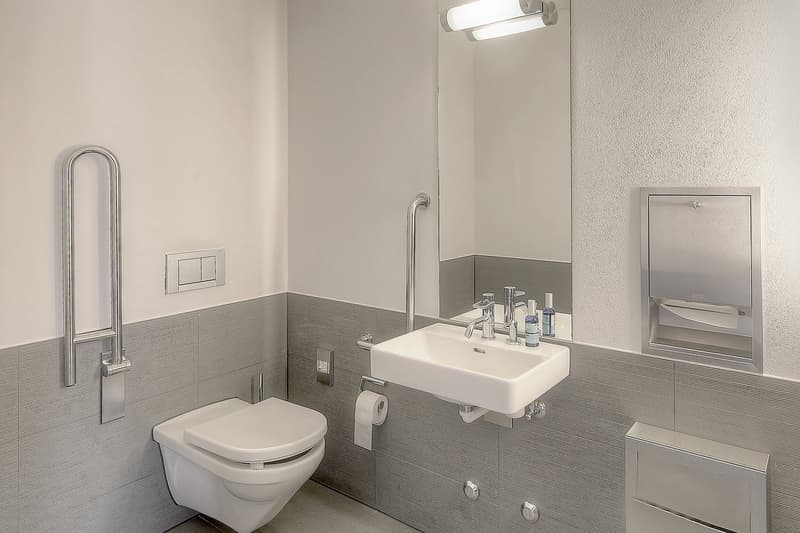 Modern bathroom with white toilet, sink, and vanity. Gray tile walls and floor. Overhead lighting and mirror. Toilet paper holder and other bathroom accessories visible.