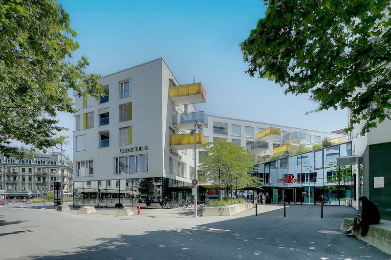 Multi-story commercial building with balconies, glass windows, and retail stores on the ground floor. The building is surrounded by trees and has a public plaza in front with benches and bicycle racks.