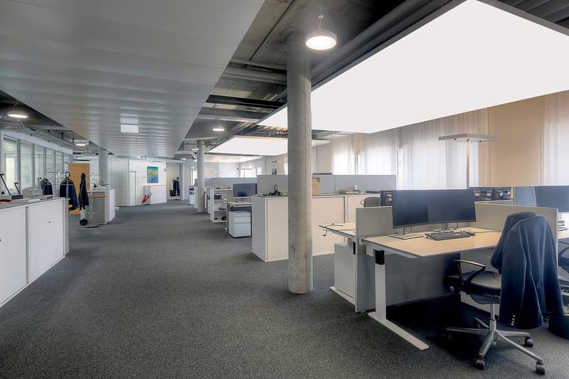 Open office layout with cubicles, desks, computers, and storage units. Exposed ceiling with lighting fixtures. Carpeted floor. Concrete columns visible.