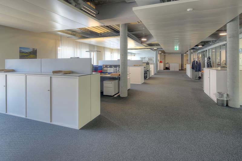 Large open office space with cubicles, storage cabinets, and various office equipment such as printers and copiers. The space has a modern, industrial look with exposed ceiling beams and ductwork. There is a dishwasher visible, indicating some amenities f