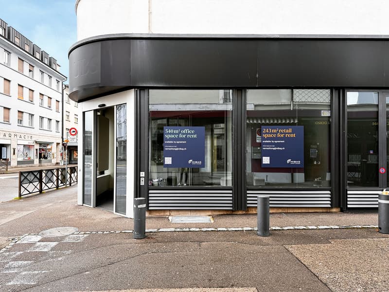Modern office building with glass front and large entrance, signage for office and retail space rental, metal bollards in front, UBS logo at the top.