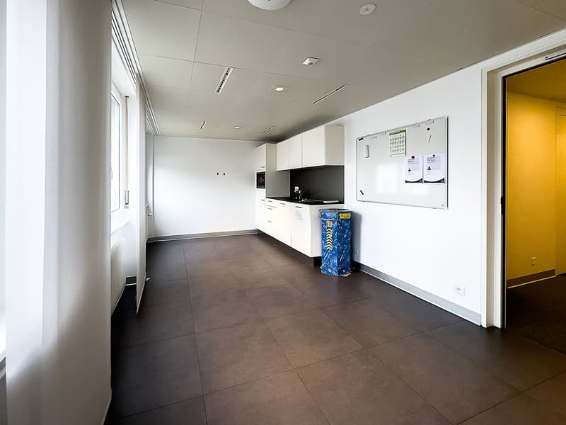 Empty room, white walls, brown floor, small kitchen area, white cabinets, microwave, sink, trash bin, bulletin board