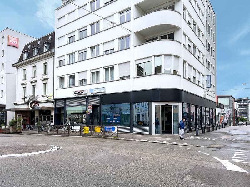 High-rise building, glass windows, black and white facade, store fronts, cafe with chairs, man walking on the sidewalk, greenery in front