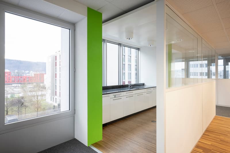 A room with large windows, wooden floors, and white walls, showcasing a city view.