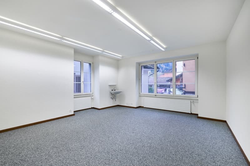 The image shows a spacious, well-lit office space with white walls, a gray carpeted floor, and large windows providing natural light. The room has a clean and minimalist design, with recessed lighting fixtures installed in the ceiling. There is a small si