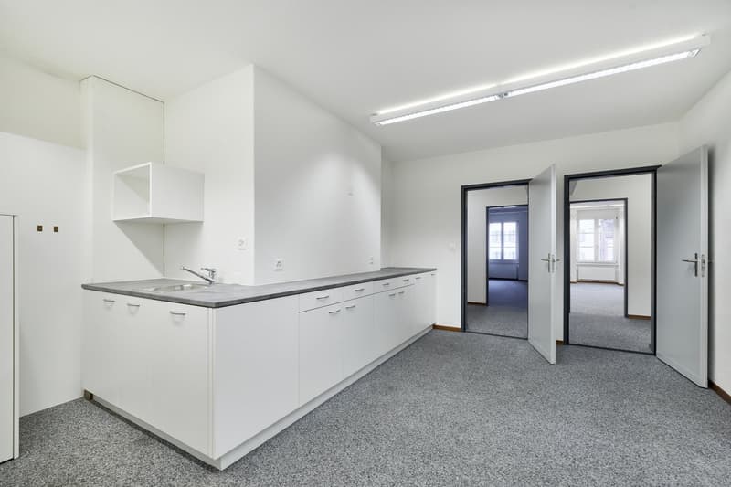 The image shows a modern, minimalist apartment interior. The main room features a white kitchen with a long counter and cabinets. The floor is covered in gray carpet, and there are two doorways leading to other rooms. The lighting is bright and the overal