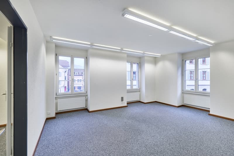 Large open office space with white walls, gray carpeted floor, and multiple windows providing natural light. The room is equipped with overhead lighting fixtures and appears to be ready for use as an office or commercial space.