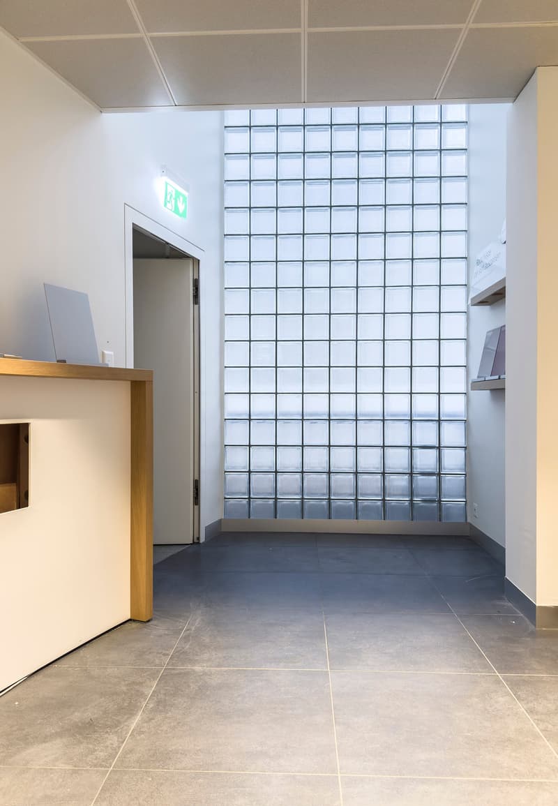 The image shows a modern office space with a large glass wall, a wooden reception desk, and a tiled floor. The space appears to be well-lit and has a minimalist, clean design.