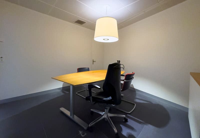 The image shows a small office space with a yellow table, black office chairs, and a pendant light fixture. The room has plain white walls and a dark floor.
