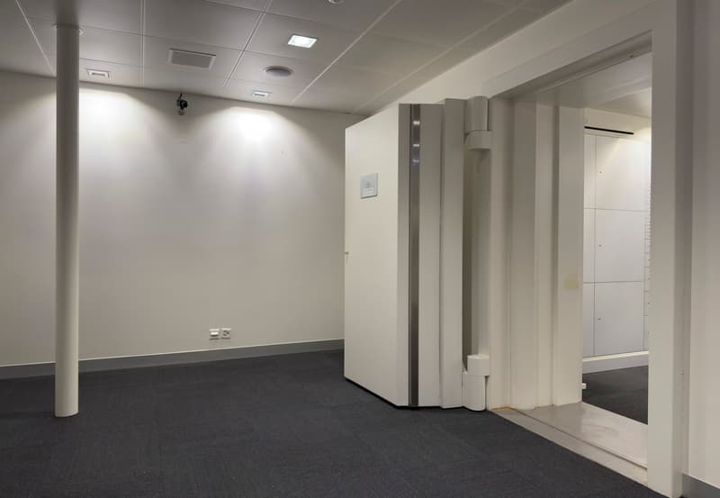 The image shows an empty office space with white walls, a gray carpeted floor, and a column in the center. There are recessed lighting fixtures in the ceiling and a thermostat on the wall. The space appears to be a commercial or office environment.