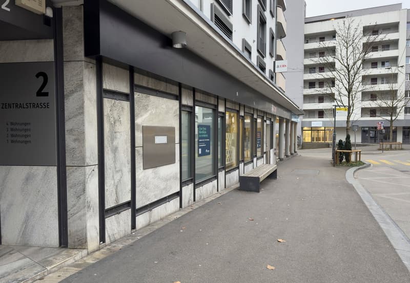 The image shows the exterior of a commercial building on Zentralstrasse. The building has a modern, minimalist design with large glass windows and a stone facade. There are several storefronts or business units along the street level, with signage indicat