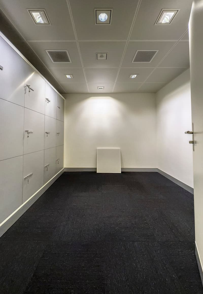 The image shows a locker room with white walls, a tiled ceiling with recessed lighting, and black carpeted flooring. There are several lockers along the walls and a small bench or table in the center of the room.
