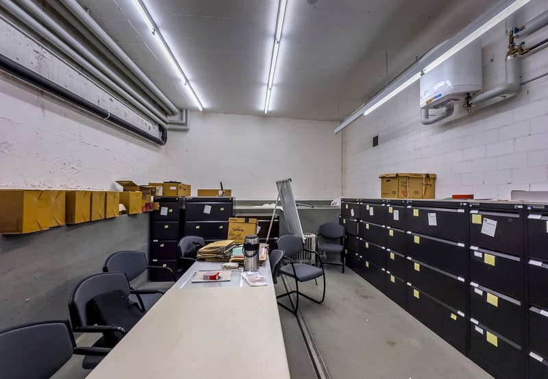 The image shows a cluttered office space with various storage boxes, filing cabinets, and office equipment. The room has a concrete floor, white walls, and overhead lighting. There is a desk with a computer monitor and other office supplies, as well as se