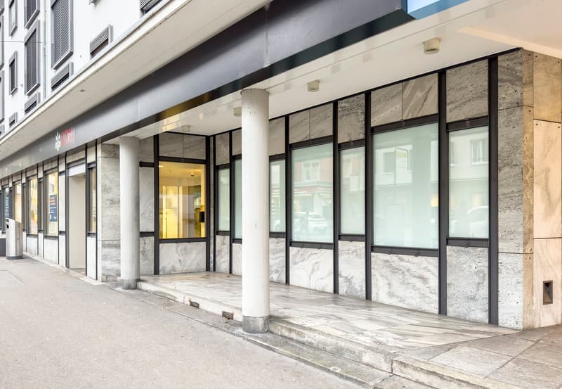 The image shows the exterior of a commercial building with a modern, minimalist design. The building has a glass and metal facade with large windows, and the entrance is framed by stone columns. The overall aesthetic is sleek and contemporary.