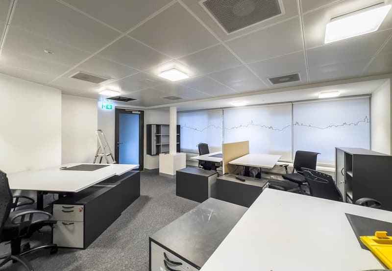 The image shows a modern office space with a large open floor plan. The room has a gray and white color scheme, with a tiled ceiling and recessed lighting. There are several desks and workstations set up, along with storage cabinets and shelves. The walls