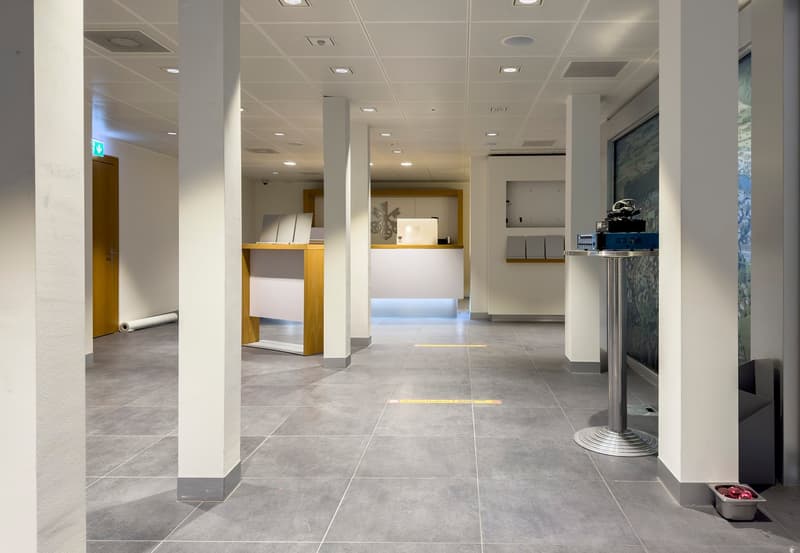 Large open office space with gray tile flooring, white walls, and recessed lighting. There are several workstations and desks visible, as well as a reception area with a counter. The space appears to be a modern, professional office environment.