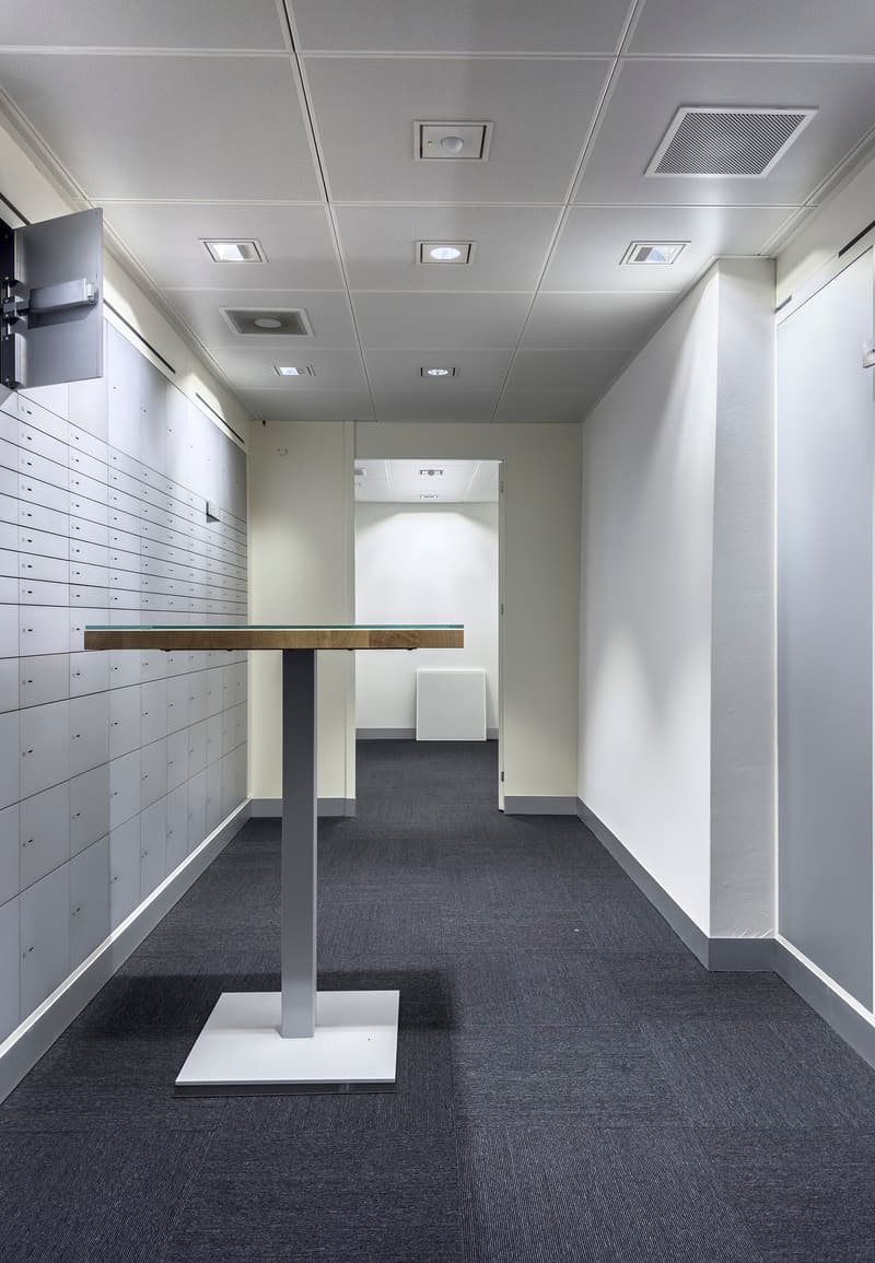 The image shows an office space with a minimalist design. The room has a gray and white color scheme, with a suspended ceiling and recessed lighting. There are rows of lockers or storage units along one wall, and a small table or counter in the center of