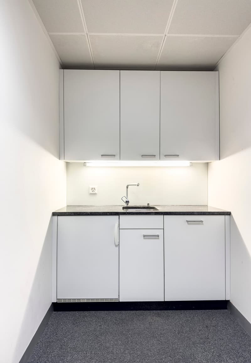 The image shows a minimalist kitchen with white cabinets, a black countertop, and a sink with a faucet. The kitchen appears to be in a small, enclosed space with a tiled floor.