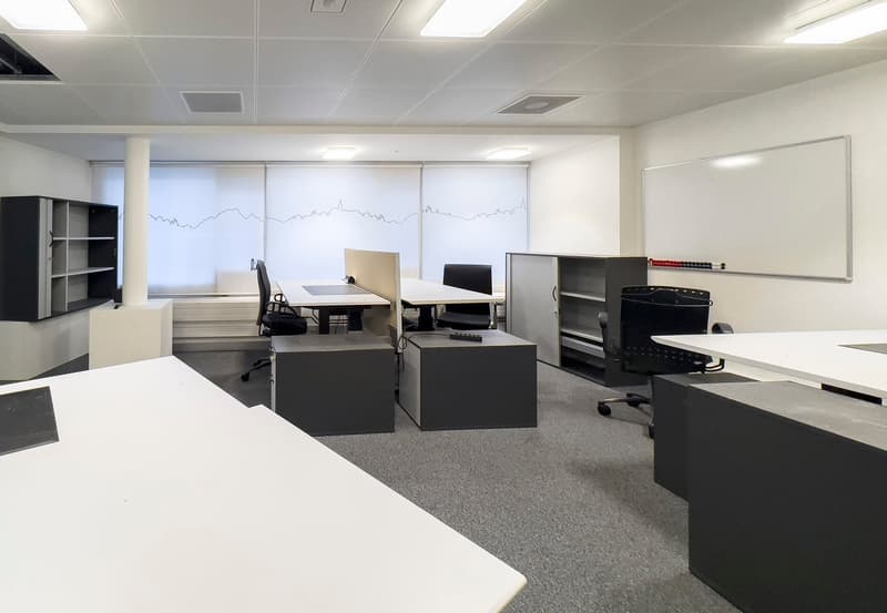 The image shows a modern office space with multiple workstations. The room has white walls, a gray carpet floor, and recessed lighting. There are several desks with computers and office chairs, as well as storage cabinets and a whiteboard on the wall.