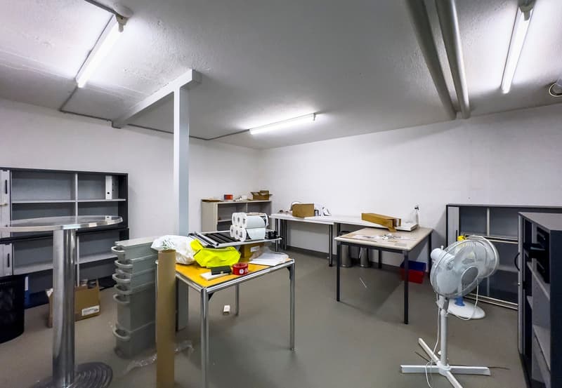 The image shows a storage room or utility space with various shelves, boxes, and equipment. The room has a utilitarian appearance with concrete floors, white walls, and overhead lighting. There are several metal shelving units, a desk or workbench, and va