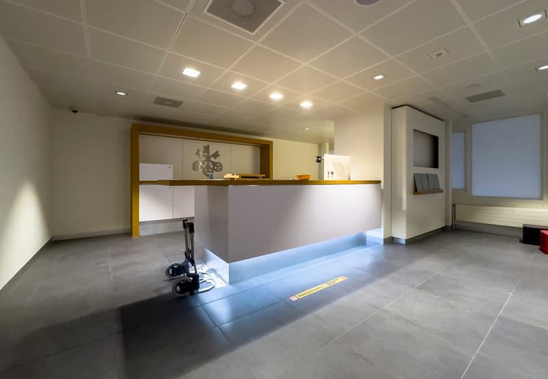 The image shows a modern office space with a reception desk area. The room has a clean, minimalist design with white walls, a gray tile floor, and a suspended ceiling with recessed lighting. The reception desk is a simple, rectangular structure with a whi