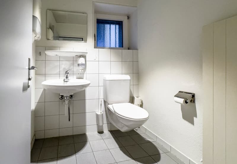 The image shows a bathroom with white tiles, a sink, a toilet, and a small window. The bathroom appears to be clean and well-maintained.