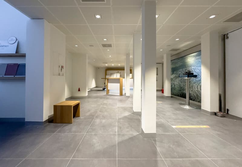 Large open office space with white walls, gray tile flooring, recessed lighting, and a wooden bench. The space appears to be a commercial or office environment.