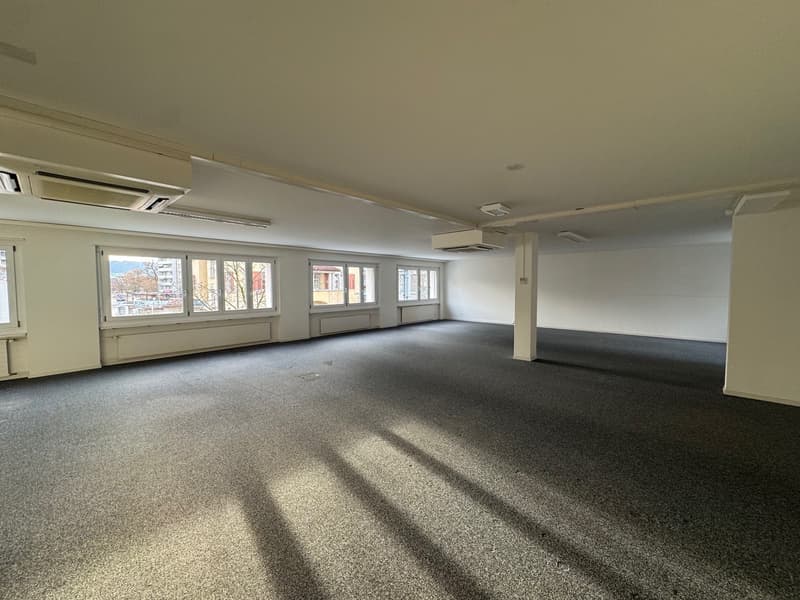 Empty room with white walls, gray carpet, large windows, and ceiling-mounted air conditioning units.