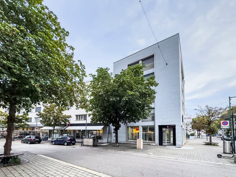 Modern building with glass windows, entrance on the street side, UBS bank sign, trees, parked cars, and a trash bin.