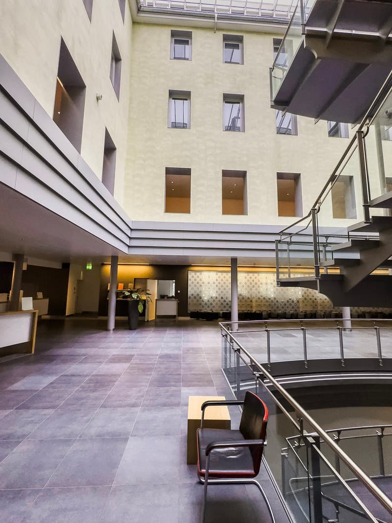 The image shows the interior of a modern building with a large open space. The floor is covered in gray tiles, and the walls are a light beige color. There are several windows on the upper floors, and the ceiling has a unique angular design. In the center