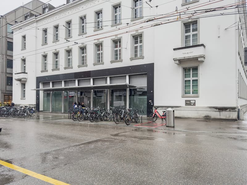 A multi-story building with a glass-fronted entrance and a bicycle parking area in front. The building has a traditional architectural style with many windows and a balcony on the upper floors.