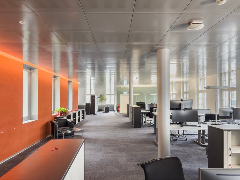 Large open office space with gray carpeted floors, white ceiling tiles, and orange accent walls. The space contains multiple workstations with computers and desks, as well as a lounge area with chairs and a table. There are also some storage units and a p