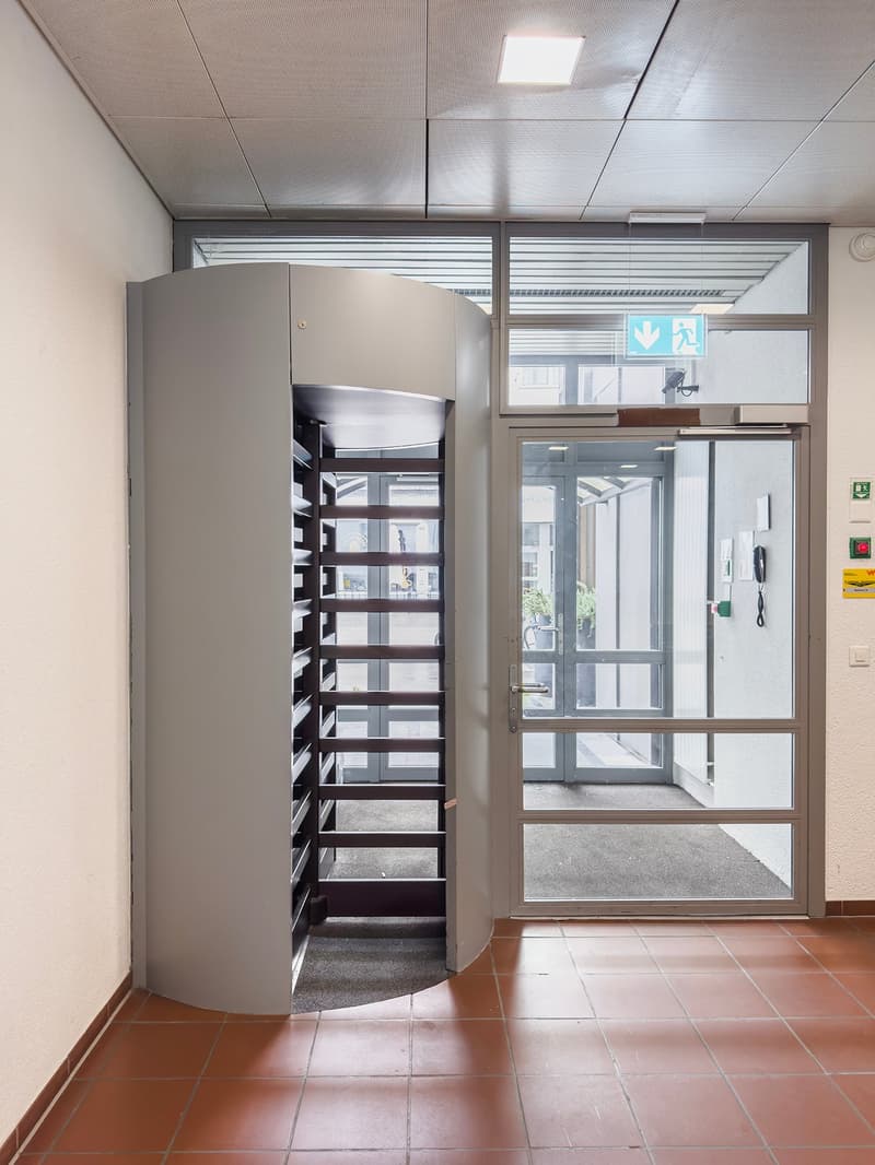 The image shows an office or commercial space with a large storage cabinet or locker, a glass entrance door, and a tiled floor. The space has a clean, modern design with a suspended ceiling and recessed lighting.