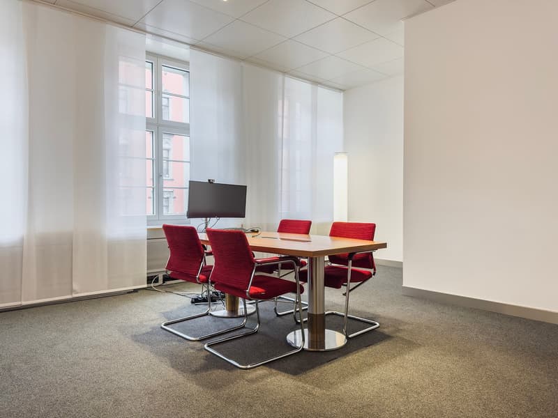 A bright and spacious conference room with large windows, a wooden table, and red upholstered chairs. The room has a TV mounted on the wall, providing a space for presentations or meetings.