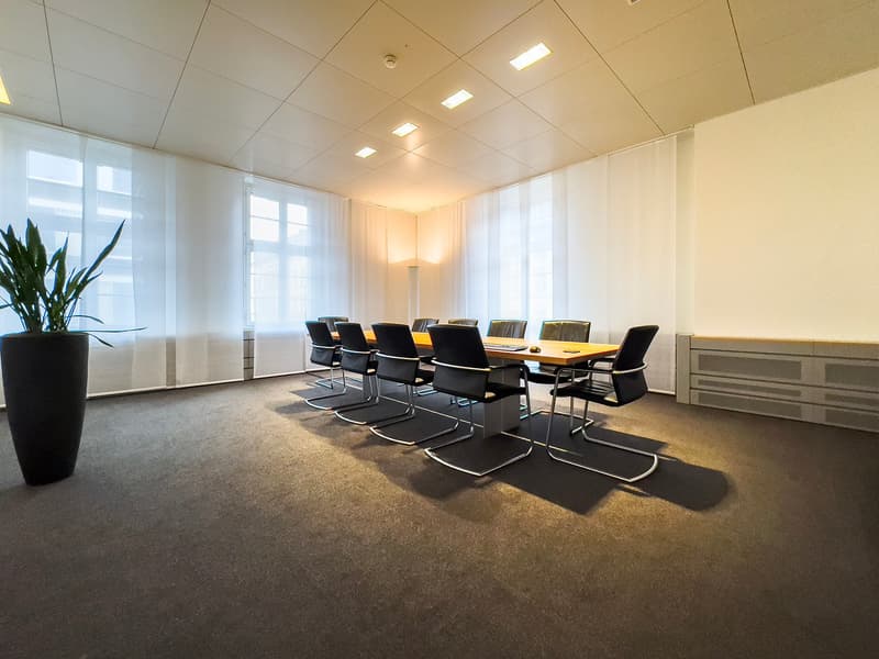 Large conference room with a long wooden table, black leather chairs, and large windows providing natural light. The room has a modern and minimalist design with recessed lighting in the ceiling.