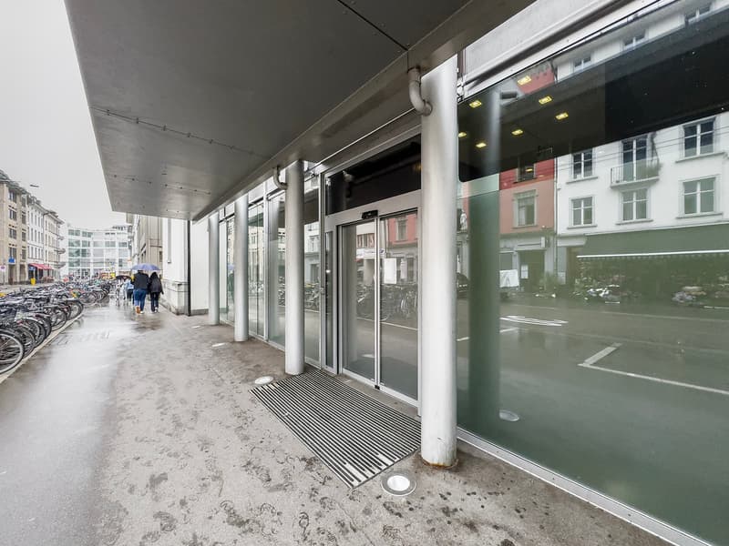 The image shows the exterior of a commercial building with glass doors and windows. There is a covered walkway with columns and a metal roof. The building has a modern, minimalist design with a gray and green color scheme. There are several bicycles parke