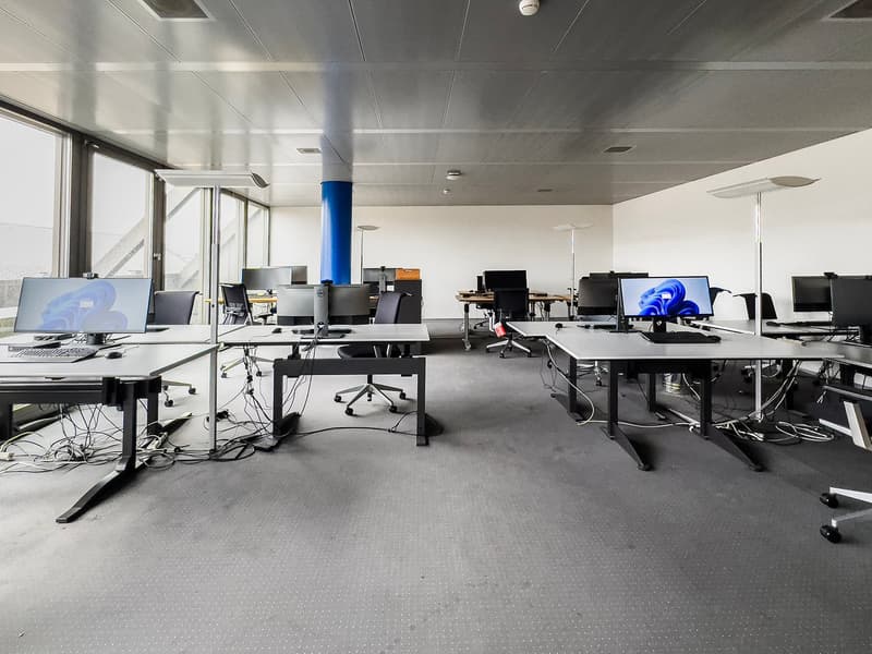 Open office space with multiple desks, computers, and office equipment. The room has a gray and white color scheme with a tiled floor and a high ceiling with recessed lighting. There is a blue column visible in the center of the room.