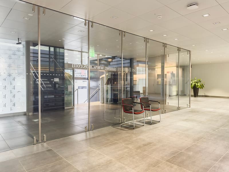 The image shows a modern, well-lit office space with glass walls and doors. The floor is covered in beige tiles, and there are several red chairs in the waiting area. The space appears to be a professional business environment, with a clean and minimalist