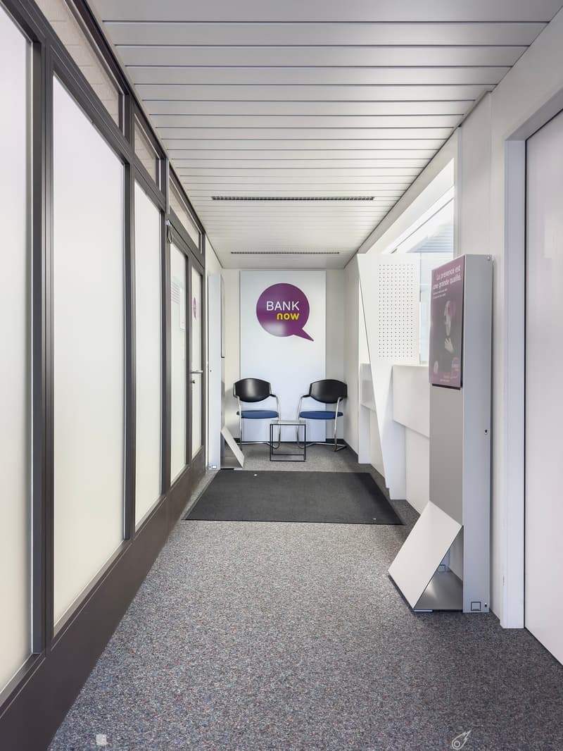 The image shows a hallway or corridor in an office space. The hallway has a gray tiled floor, white walls, and a ceiling with parallel beams. There are two black chairs placed in the hallway, and a large sign on the wall that says 'BANK now'. The hallway