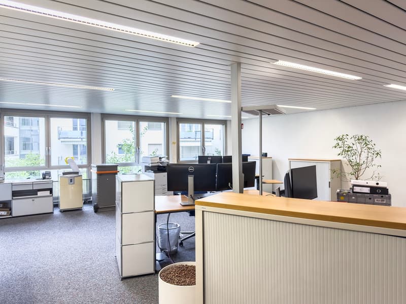 Open office space with desks, computers, and office equipment. Large windows provide natural light. Shelves and storage units are visible.