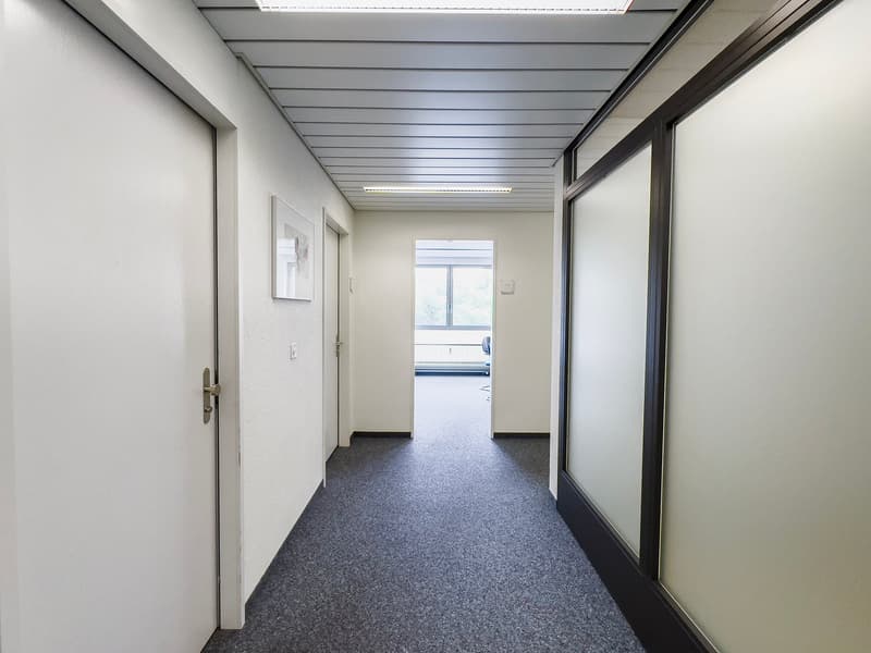 The image shows a long, narrow hallway with a gray terrazzo floor and white walls. The ceiling has a series of parallel beams running across it, creating a modern, industrial look. There are several doors lining the hallway, suggesting this is an office o