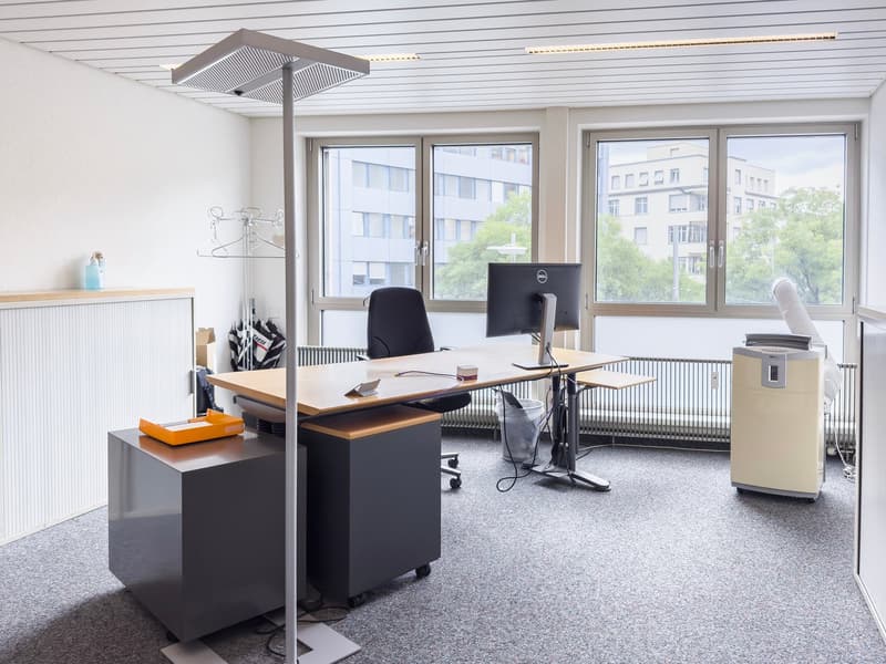 Modern office space with large windows, desk, computer, and office equipment. The room has a minimalist and clean design with white walls and gray flooring.